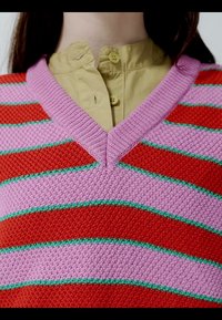 Gros plan sur une personne portant une chemise beige boutonnée sous un pull en maille rayé rose, rouge et vert avec un col en V.
