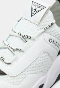 Bela superga z zgornjim delom iz mrežastega materiala in teksturiranimi usnjenimi poudarki. V ospredju je črn napis "GUESS" in ravni beli vezalke.