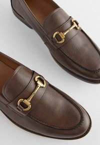 Mocassins en cuir marron avec une finition texturée, accents en métal doré, et un design à bout rond avec des détails de couture contrastants.