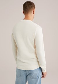 Weißer strukturierter Pullover mit Rautenmuster, Rundhals und gerippten Bündchen. Kombiniert mit hellblauen Jeans.