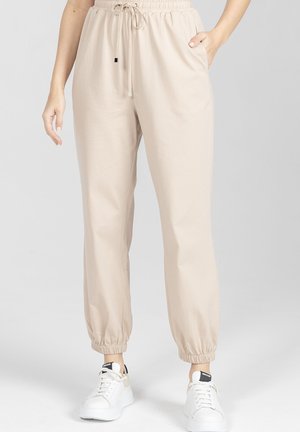 Beżowe spodnie dresowe z elastycznym pasem i ściągaczami przy kostkach, noszone z białymi sneakersami na platformie, jedna ręka w kieszeni, neutralne tło.