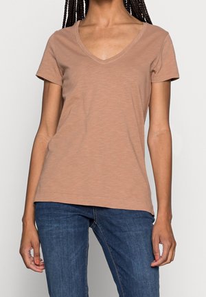 Kobieta ubrana w gładki brązowy t-shirt z krótkim rękawem i dekoltem w serek oraz niebieskie dżinsy, stojąca z rękami swobodnie opuszczonymi wzdłuż ciała.