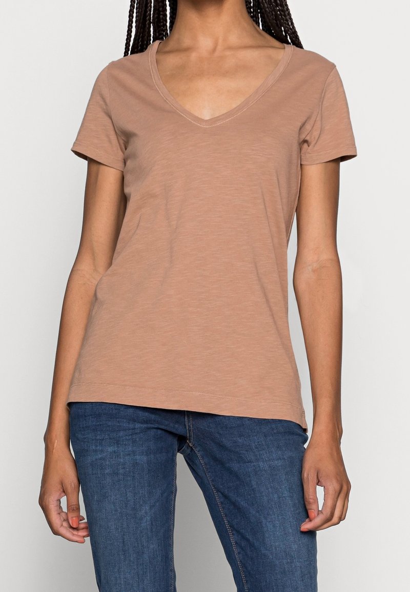 Femme portant un t-shirt marron uni à col en V et manches courtes, ainsi qu'un jean bleu, debout avec les bras détendus le long du corps.