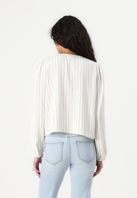 White long-sleeve blouse with vertical black stripes, slightly boxy fit, gathered sleeves, and a cropped hemline, paired with light blue jeans.