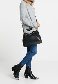 Sort håndtaske lavet af glat materiale med gyldent hardware, struktureret form og top håndtag, ledsaget af en grå sweater og denim jeans.