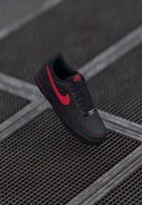 Black leather Nike Air Force 1 sneakers with red accents, featuring a perforated toe, black laces, and a textured rubber sole.