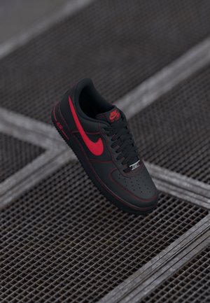 Black leather Nike Air Force 1 sneakers with red accents, featuring a perforated toe, black laces, and a textured rubber sole.