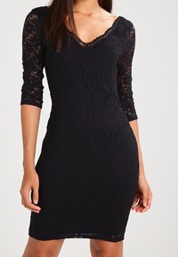 Frau in einem figurbetonten schwarzen Spitzenkleid mit Dreiviertelärmeln und V-Ausschnitt, die vor einem schlichten Hintergrund steht.