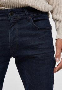 Tmavě modré džíny z denimu mají úzký střih, klasický design s pěti kapsami a jemné švy podél švů a kapes.