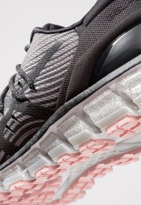 Gros plan d'une chaussure de course texturée noire et grise avec lacets, mettant en valeur une semelle transparente avec une bande de roulement rose et un support de talon noir.