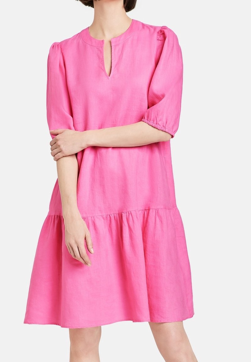 Femme portant une robe ample rose vif avec des manches bouffantes mi-longues et une petite fente à l'encolure devant, sur un fond uni.