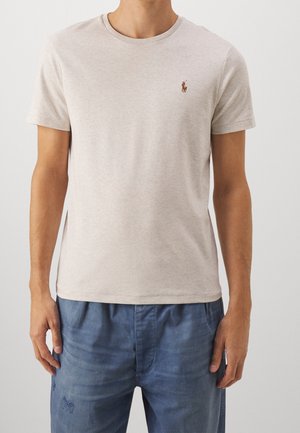 Homme portant un t-shirt beige clair à manches courtes avec un petit logo brodé sur la poitrine et un pantalon décontracté bleu.