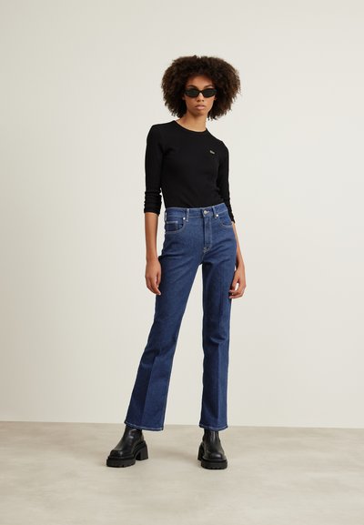 Haut noir à manches longues avec un petit logo vert, associé à un jean flare bleu foncé et des bottes noires à talons épais. Fond blanc.