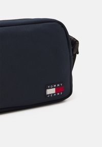 Mørkeblå rektangulær bag laget av teksturert stoff, med et fremtredende brodert "TOMMY JEANS" logo-patch i rødt, hvitt og blått.