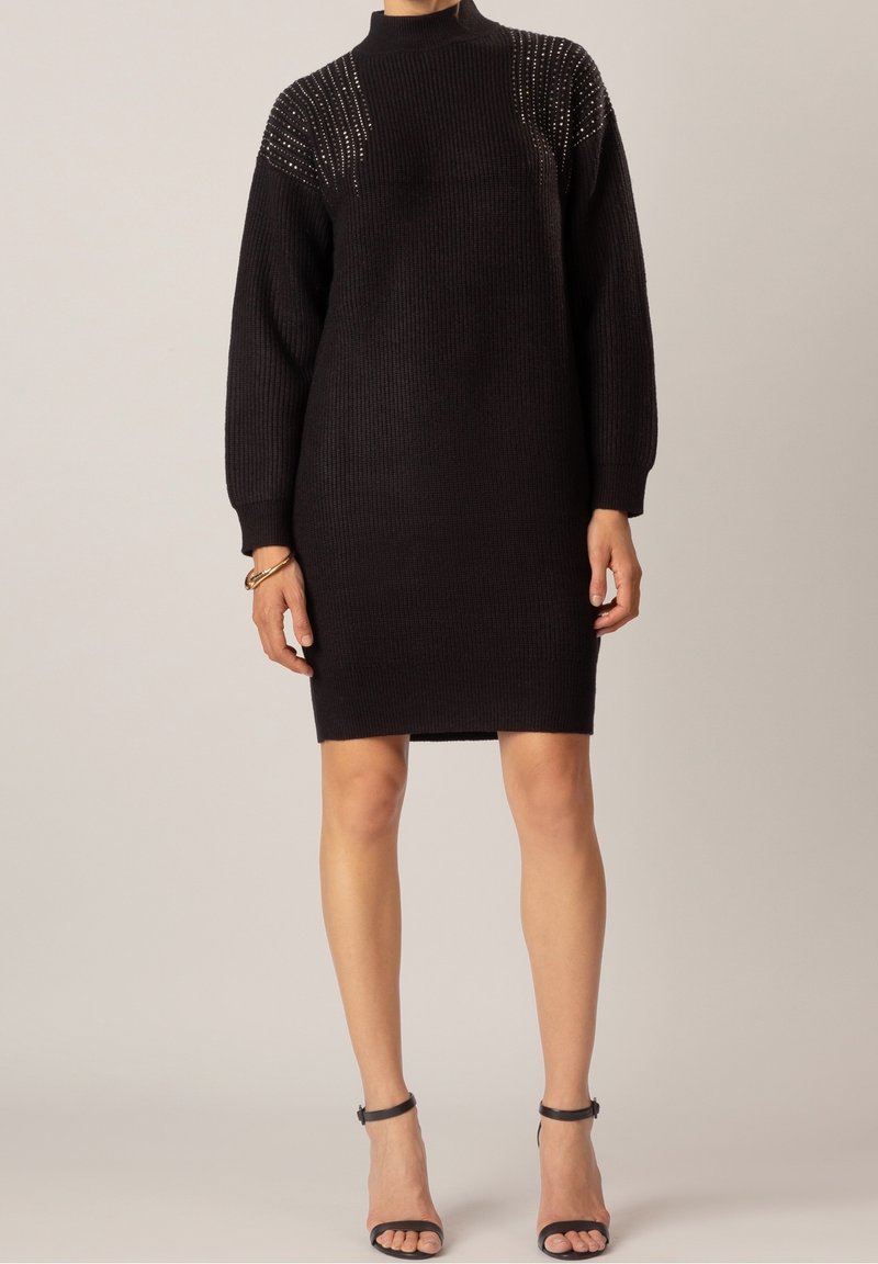 Robe noire tricotée avec un col montant, présentant un tissu texturé et des épaules ornées. Mise en valeur par une silhouette ajustée.