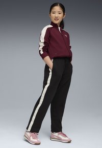 Giacca a track bordeaux con strisce bianche, pantaloni neri con pannelli laterali bianchi e scarpe rosa con dettagli bianchi. Modella in posa con le mani nelle tasche.