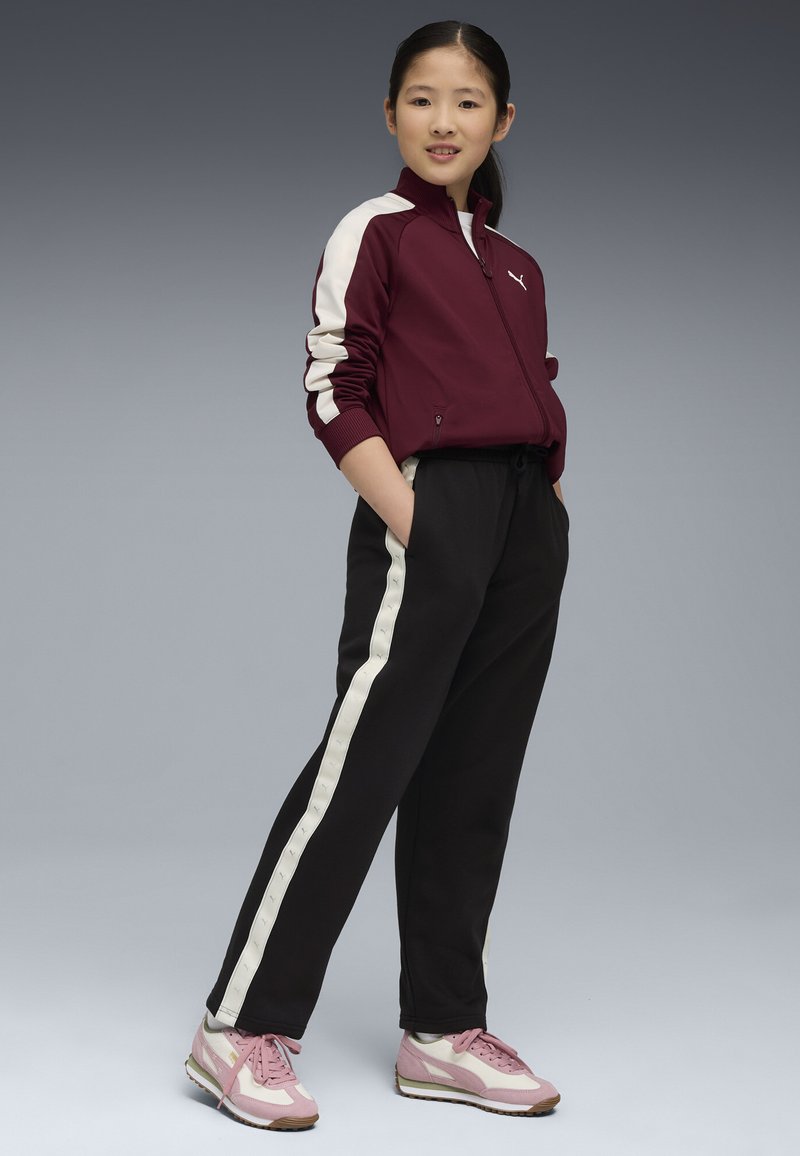 Giacca a track bordeaux con strisce bianche, pantaloni neri con pannelli laterali bianchi e scarpe rosa con dettagli bianchi. Modella in posa con le mani nelle tasche.