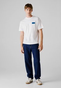 Vit bomullst-shirt med korta ärmar, med en blå rektangulär logotyp. Matchas med mörkblå jeans av denim och casual sneakers.