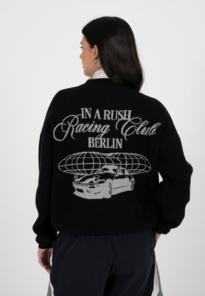 Zwarte gebreide trui met een grafisch ontwerp van een auto en een wereldbol op de achterkant, met witte tekst: "IN A RUSH Racing Club BERLIN."