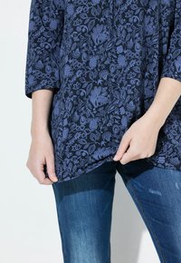 Tunique à motifs floraux bleu marine avec des manches trois-quarts, associée à un jean en denim foncé. La tunique a une coupe décontractée et une texture douce.