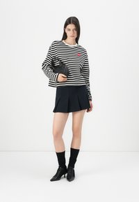 Donna che indossa un top a maniche lunghe bianco e nero a righe e una gonna plissettata nera, con una borsa nera, in piedi contro uno sfondo bianco.