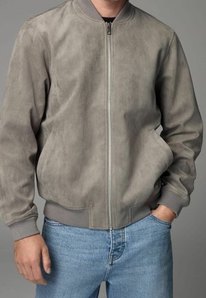 Homme portant une veste bomber en suède gris clair avec fermeture éclair à l'avant et poignets côtelés, associée à un jean bleu clair sur un fond gris uni.