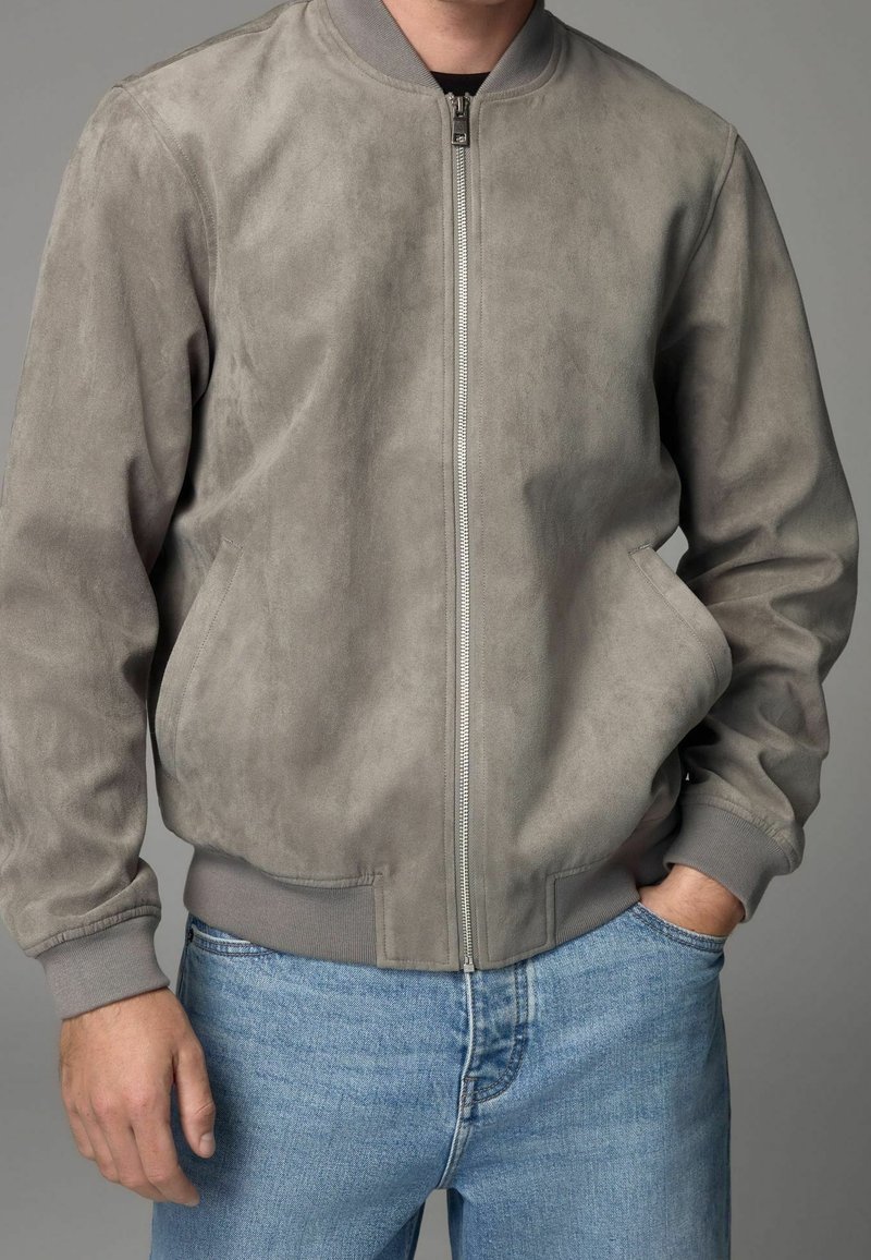 Homme portant une veste bomber en suède gris clair avec fermeture éclair à l'avant et poignets côtelés, associée à un jean bleu clair sur un fond gris uni.