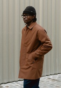 Hombre con gorro negro, gafas negras y abrigo marrón con las manos en los bolsillos, de pie frente a una pared de metal perforado.