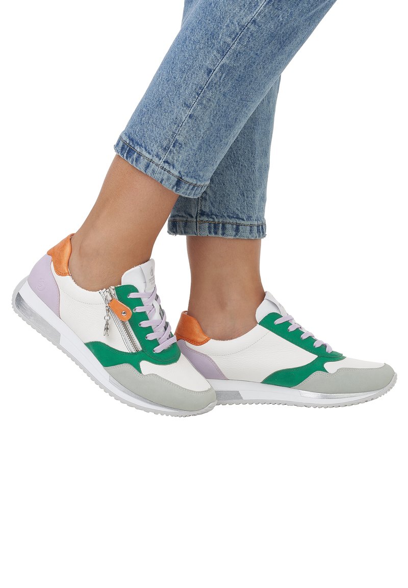 Personne portant un jean bleu et des baskets blanches avec des accents verts, orange et gris, avec une fermeture éclair latérale et des lacets.