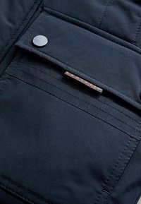 Tissu de veste rembourrée bleu marine avec une poche cousue, dotée d'un bouton-pression en argent et d'une étiquette noire avec des lettres rouges.