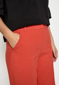 Rote maßgeschneiderte Hose mit glatter Textur, ausgestattet mit Seitentaschen und geradem Bein Design. Getragen mit einem schwarzen, locker sitzenden Oberteil.