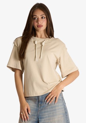 Giovane donna con lunghi capelli castani che indossa una maglietta beige a maniche corte con coulisse e jeans di denim azzurro chiaro, in piedi con una mano sui fianchi.