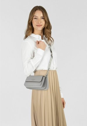 Sac bandoulière en cuir gris avec un design superposé, une chaîne argentée et une texture lisse, porté par une personne en chemise blanche et jupe beige.