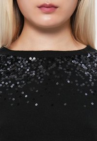 Gros plan sur la partie inférieure du visage et le cou d'une personne portant un pull noir décoré de sequins noirs et argentés dispersés près de l'encolure.