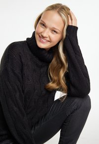 Schwarzer Kabelstrickpullover mit hohem Stehkragen und gerippten Akzenten. Das Model trägt dunkle Jeans. Glattes, strukturiertes Material.