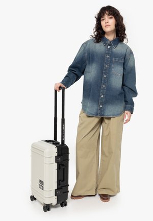 Hard-shell suitcase in off-white and black with a textured finish, dual wheels, and a retractable handle, paired with denim shirt and beige pants.