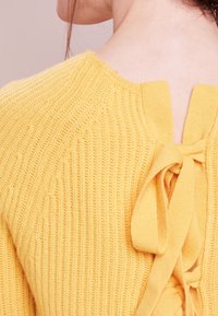 Gelbes gestricktes Sweatshirt mit strukturiertem Muster, geripptem Design und offenem Rücken mit Bindebändern. Weicher Stoff mit entspannter Passform.