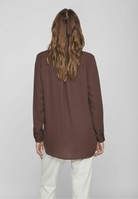 Brun långärmad skjorta med rak skärning, har en söm bak och knappade manschetter, kombinerad med vita byxor, visad bakifrån.