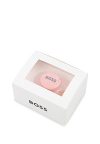 Ροζ πιπίλα σε διαφανή συσκευασία, με κυκλική ασπίδα που φέρει τη λέξη "BOSS" σε μαύρο, περιτριγυρισμένη από λευκό χάρτινο κουτί.