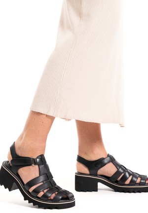 Black leather sandals with open-toe design and crisscross straps. Chunky heel with jagged edge and adjustable ankle strap.