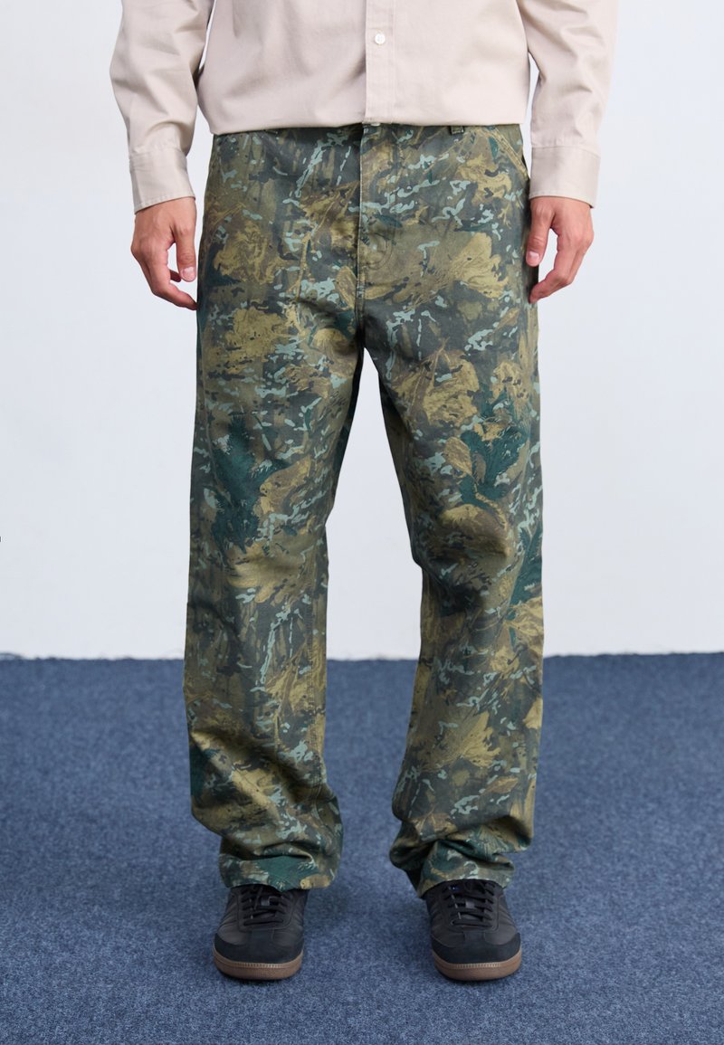Pantaloni cargo con motivo mimetico in verde e khaki, caratterizzati da una vestibilità ampia, tessuto resistente e tasche laterali per la praticità. Abbinati a sneakers nere.