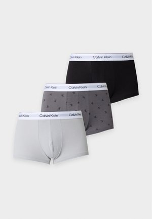 Gespierde man staat op blote voeten in witte Calvin Klein boxershorts tegen een effen lichte achtergrond, met één arm gekruist en rustend op zijn torso.