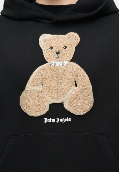 Beige teddybeerpatch met detail van witte metalen halsband op zwarte hoodie, tekst "Palm Angels" onder de beer.