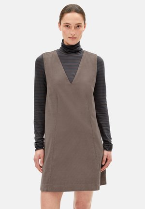 Bruine corduroy jurk met een diepe V-hals, gedragen over een zwart-grijs gestreepte turtleneck. Gladde textuur, mouwloos ontwerp.