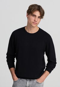 Maglione nero in una texture morbida con scollatura rotonda, polsini e orlo a coste. Indossato su pantaloni in denim grigio chiaro. Design casual.