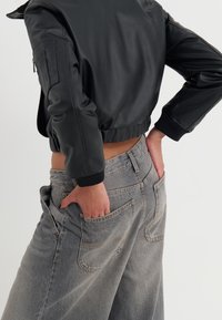 Chaqueta de cuero negra corta con puños acanalados, combinada con pantalones de mezclilla de pierna ancha de tiro alto en gris claro que tienen bolsillos traseros.