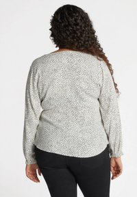 Langärmlige Bluse aus weißem Stoff mit einem schwarz gesprenkelten Muster, mit lockerer Passform und gerafften Manschetten. Abgerundeter Saum hinten.