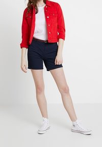 Röd denimjacka, vit t-shirt, marinblå shorts med bälte och vita högsnörda sneakers. Släta texturer, avslappnad passform och enkel design.