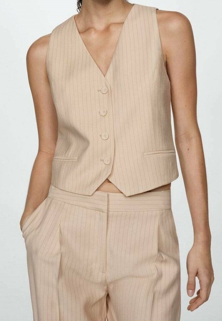 Femme portant un gilet sans manches à rayures beige avec des boutons et un pantalon taille haute assorti, une main dans la poche, sur fond uni.