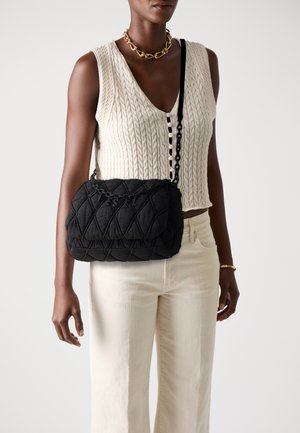 Black quilted handbag with a textured surface, featuring a chain strap. Worn over a cream knitted top and paired with light-colored pants.